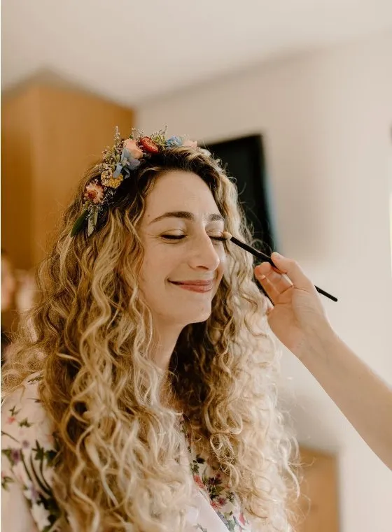 A woman with long, curly blonde hair wearing a floral headband, having her makeup applied by a makeup artist.