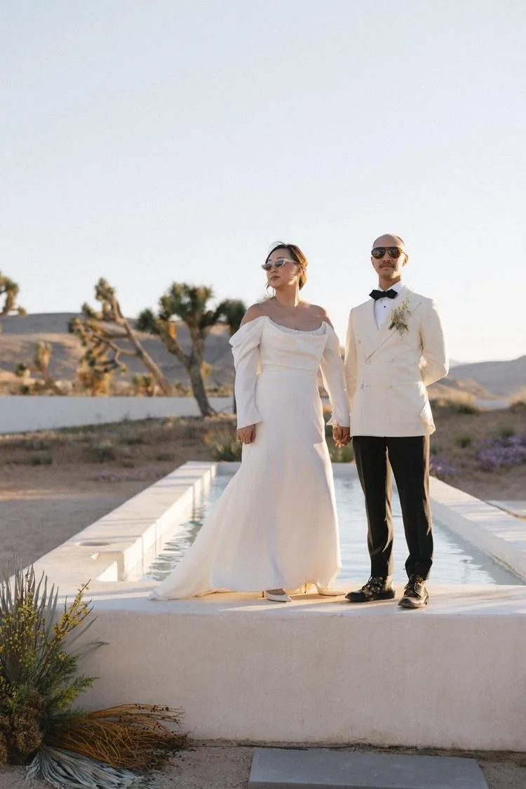 A couple dressed in wedding attire stands holding hands in an outdoor desert setting with a fountain behind them, during sunset.