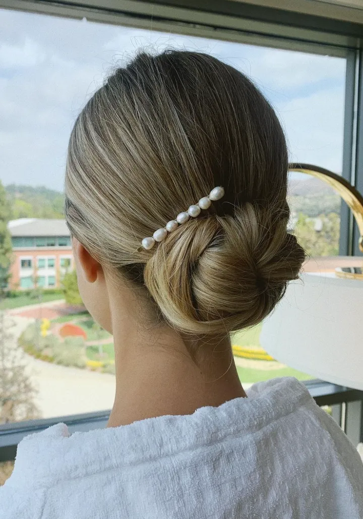 Back view of a woman with blonde hair styled in a low bun, adorned with a pearl hairpin, looking out a window at a scenic outdoor landscape.