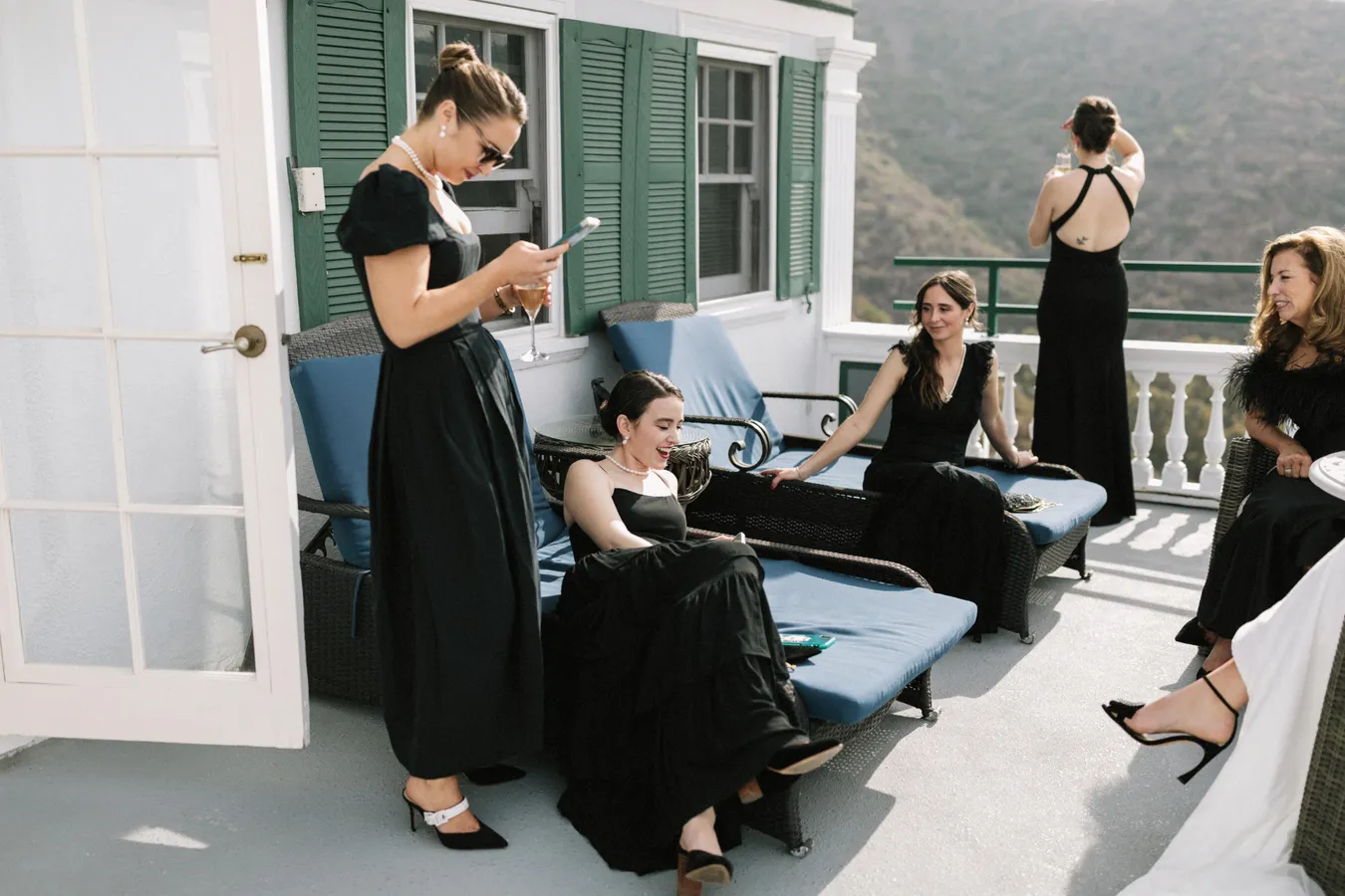 Five women dressed in black evening gowns are on a balcony with a mountain view. One woman