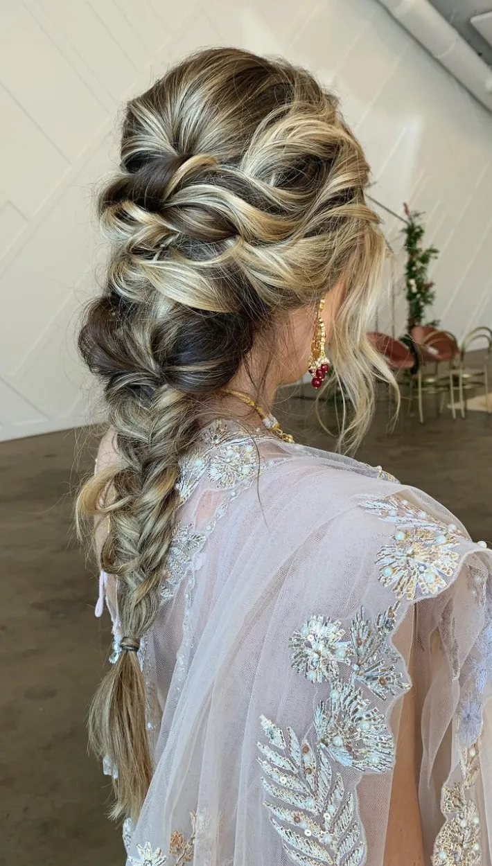 A bride with a beautifully styled updo hairstyle featuring a side braid and loose curls, wearing elegant earrings and a sheer embroidered dress.