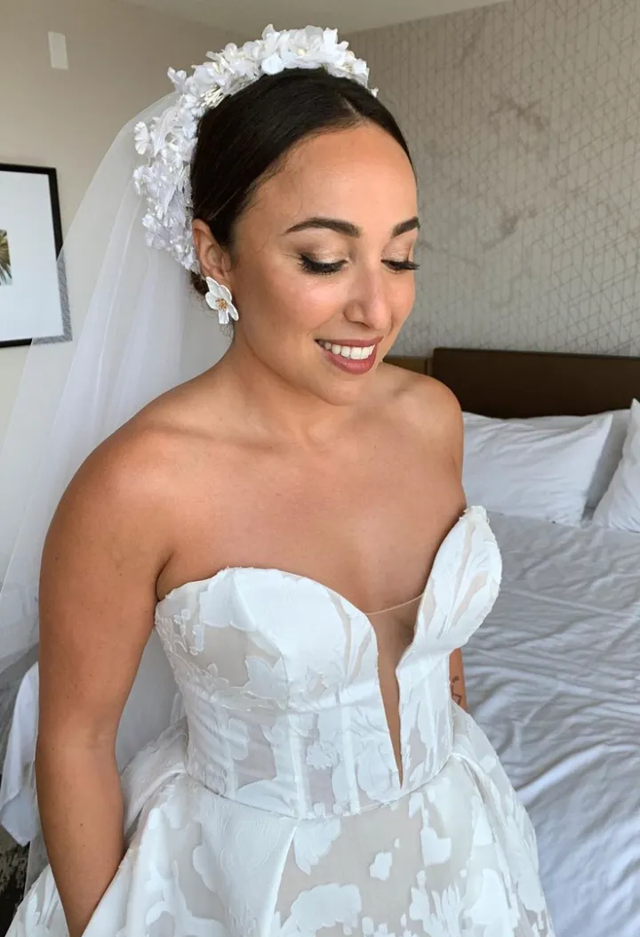 A bride with dark hair wearing a white strapless wedding dress with floral details, a white floral headpiece, and white flower earrings, smiling with eyes closed, standing in a bedroom.