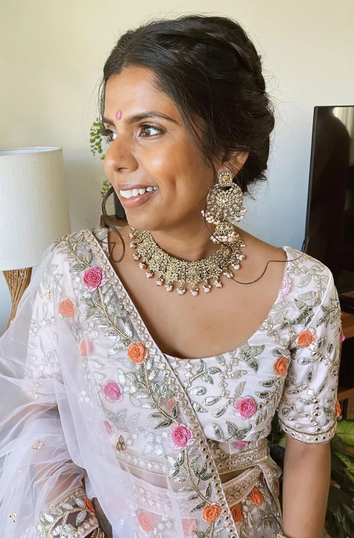 A bride dressed in traditional Indian attire, wearing intricate jewelry and a embroidered saree with floral patterns, smiling and looking to her left.