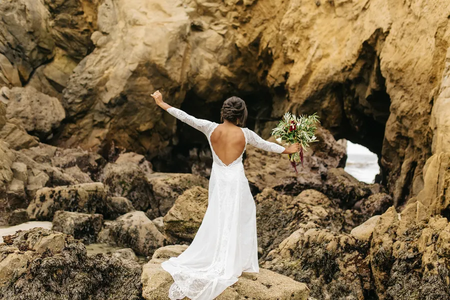 A woman in a white wedding dress stands on rocks in front of a natural rock arch formation, holding a bouquet of flowers with her arms outstretched in big sur