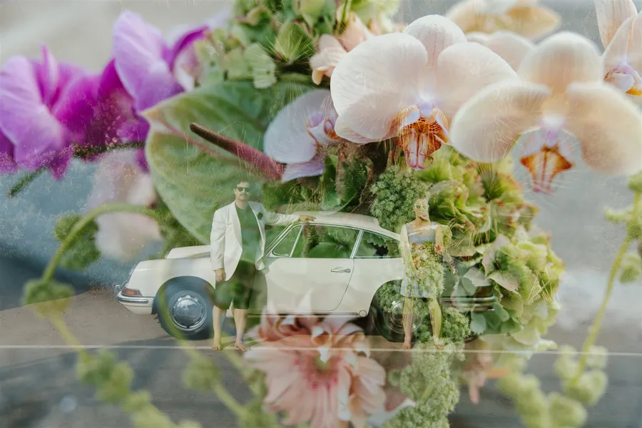 Bride and groom standing beside a vintage white car on a road, overlaid with a large, colorful floral image.