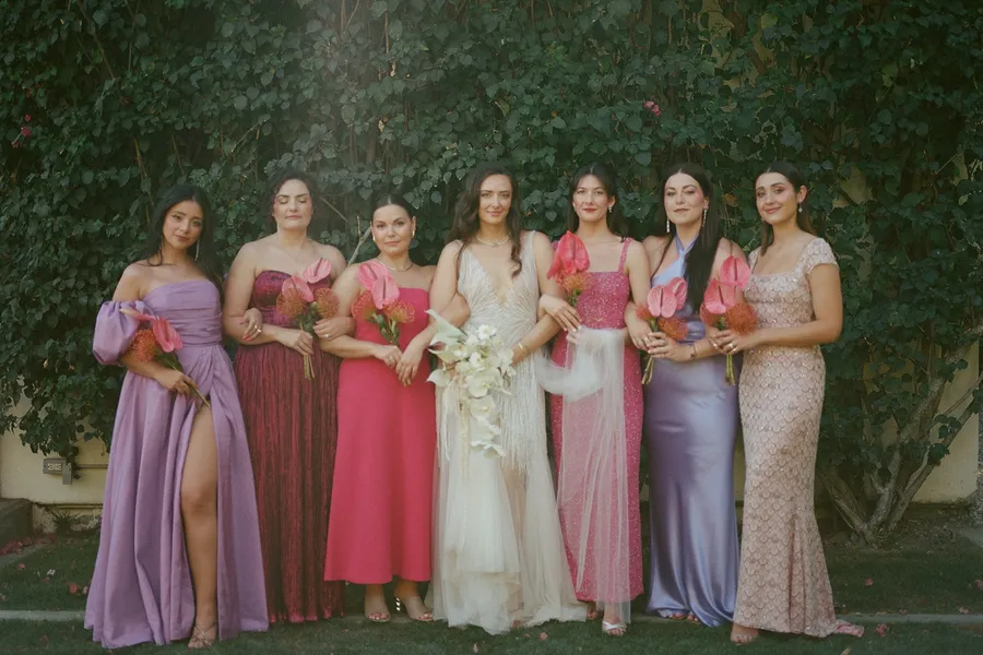 A bride in a white wedding dress standing with seven women in colorful dresses, all holding bouquets of pink flowers, outdoors in front of a green hedge at The Parker Palm Springs.