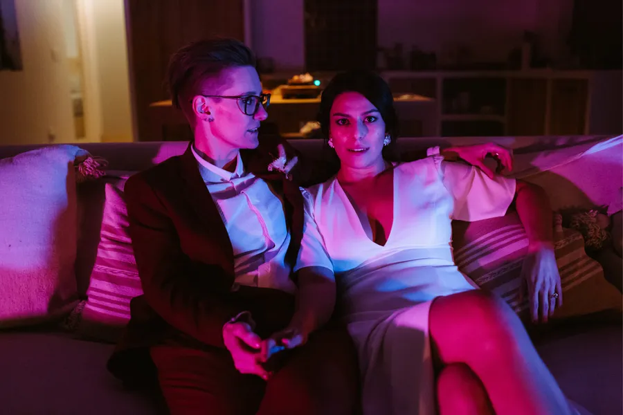 Two brides sitting on a sofa in a dimly lit room with pink and purple lighting. The brides are wearing glasses and a suit, holding a remote and one is dressed in a white dress with floral earrings, looking towards the camera.