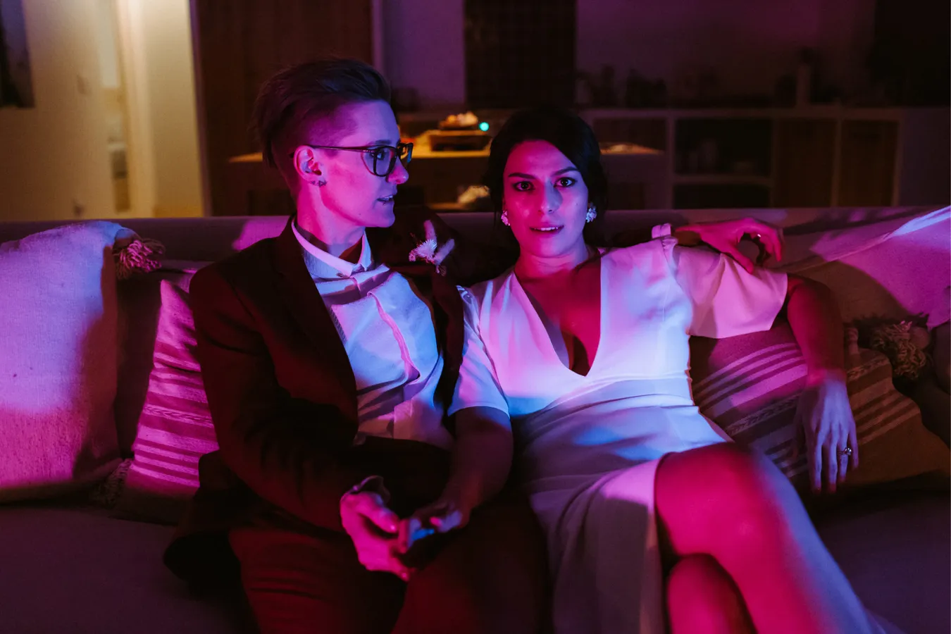 A couple sitting closely on a sofa in a dimly lit room with pink and purple lighting. One