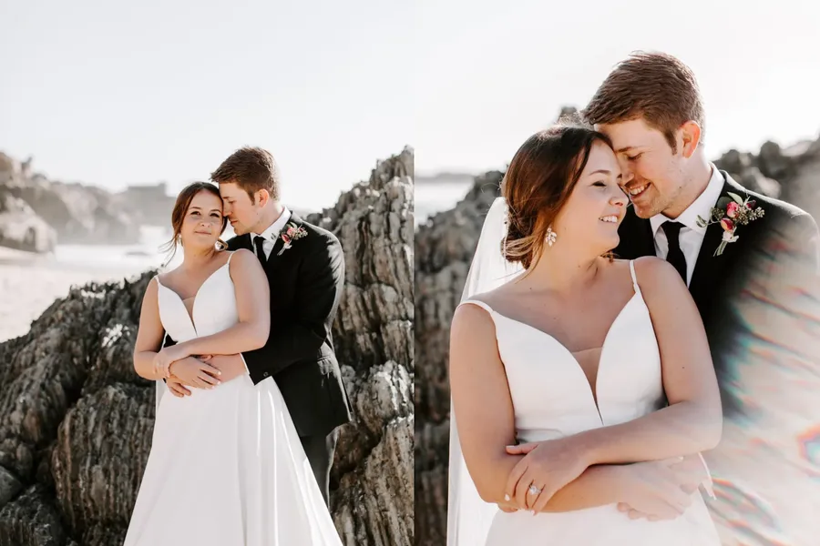 A bride and groom on the beach, standing close together, with rocky formations in the background. The bride is wearing a white wedding dress, and the groom is in a black suit with a tie and boutonniere. They are smiling and leaning their foreheads to