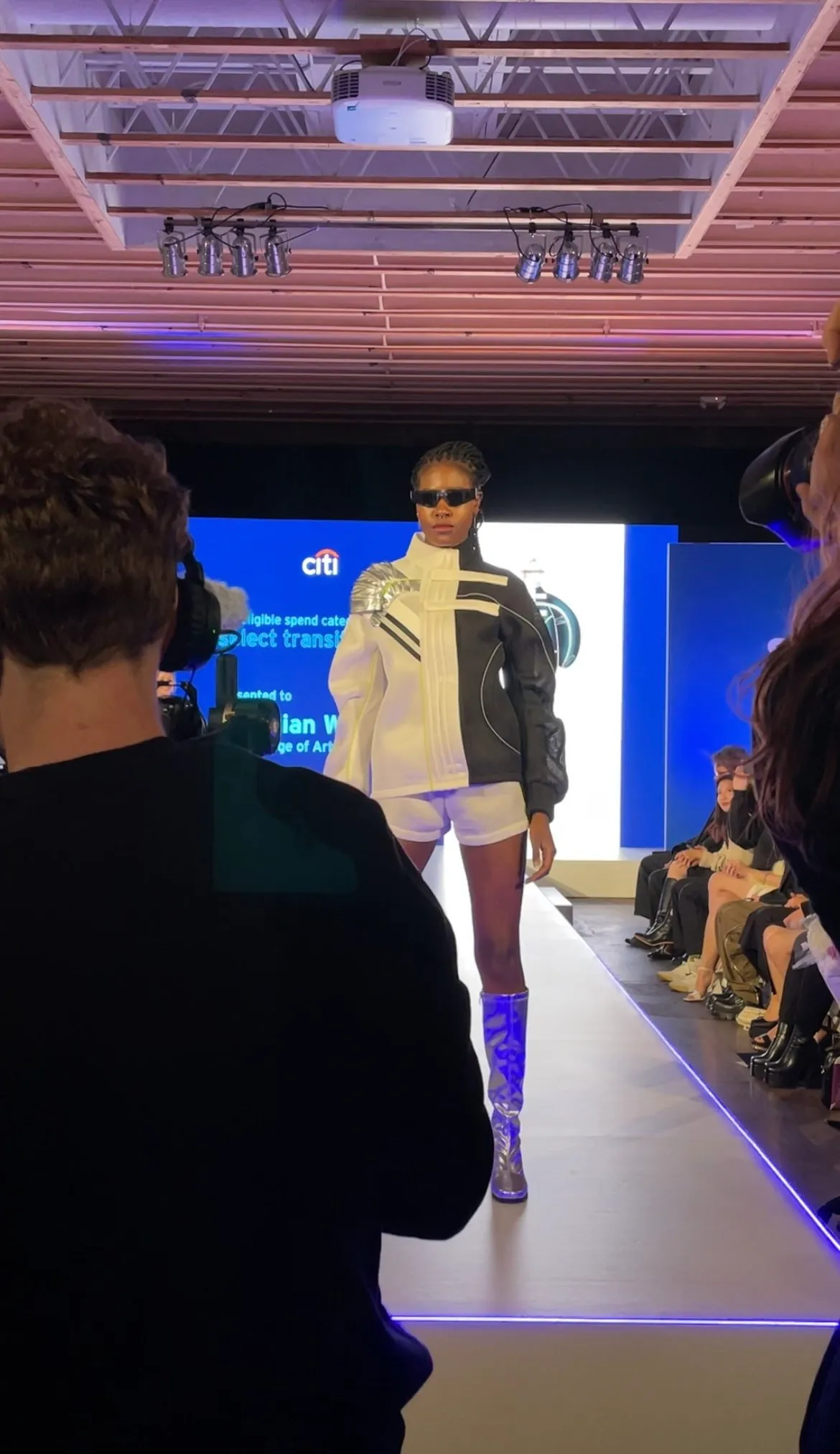A fashion show runway with a model walking in a white and black futuristic jacket, white shorts, and knee-high silver boots, wearing sunglasses, with an audience seated on either side and a large blue digital screen behind her.