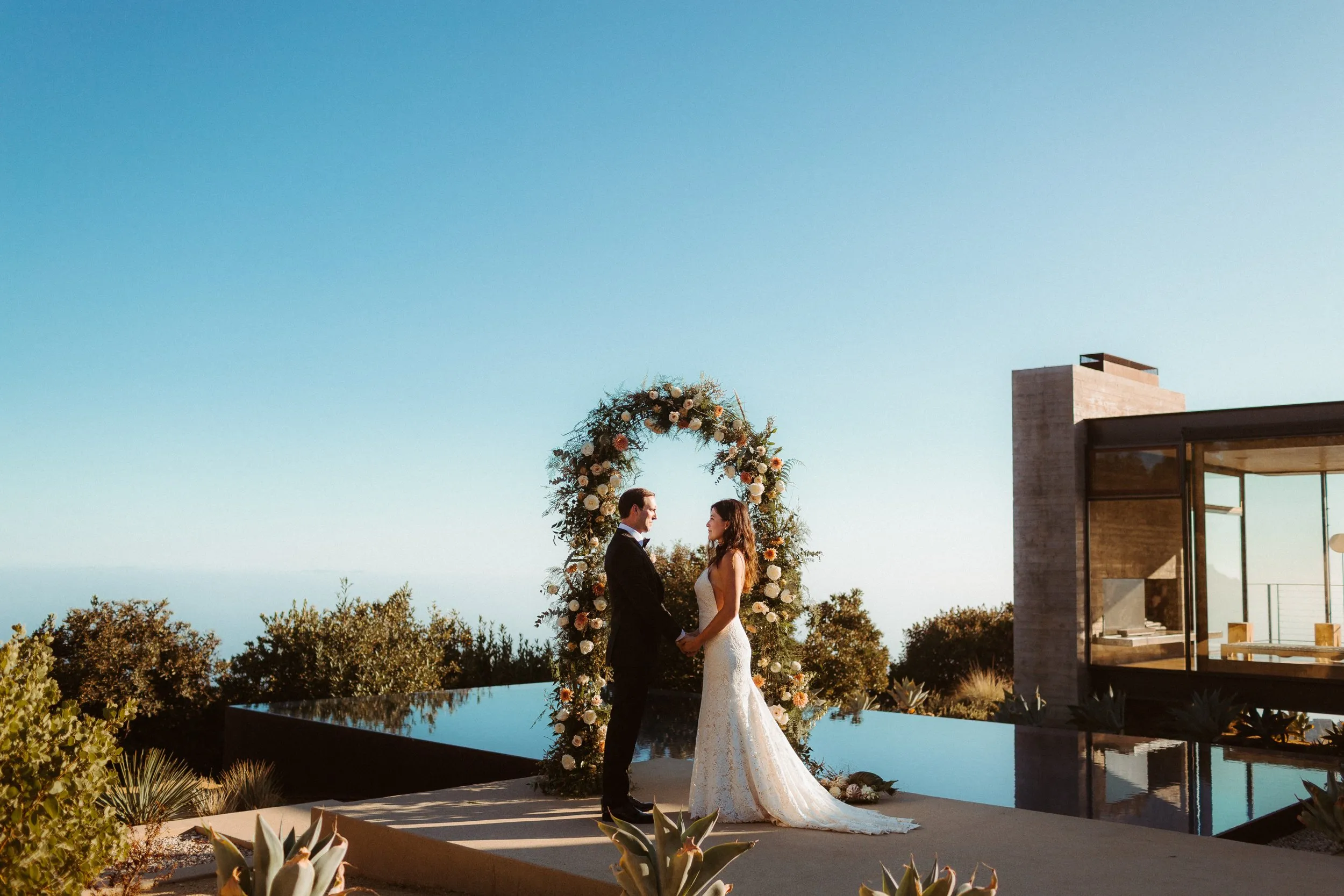 Bride and groom holding hands under a floral arch beside an infinity pool at a modern hilltop venue