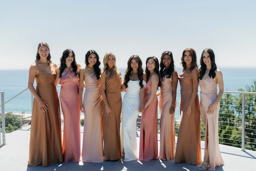 Group of ten women in evening gowns standing outdoors on a balcony with a view of the ocean in the background at a malibu wedding