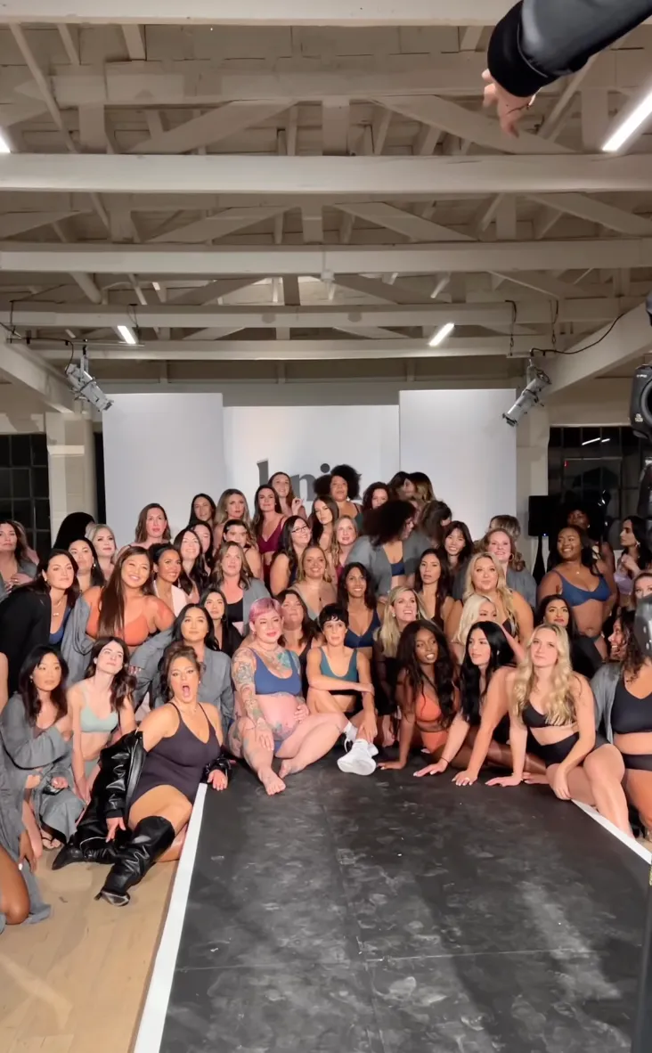 Group of women gathered backstage at a fashion show or event, posing for a photo, with a runway in front of them and a white backdrop behind.