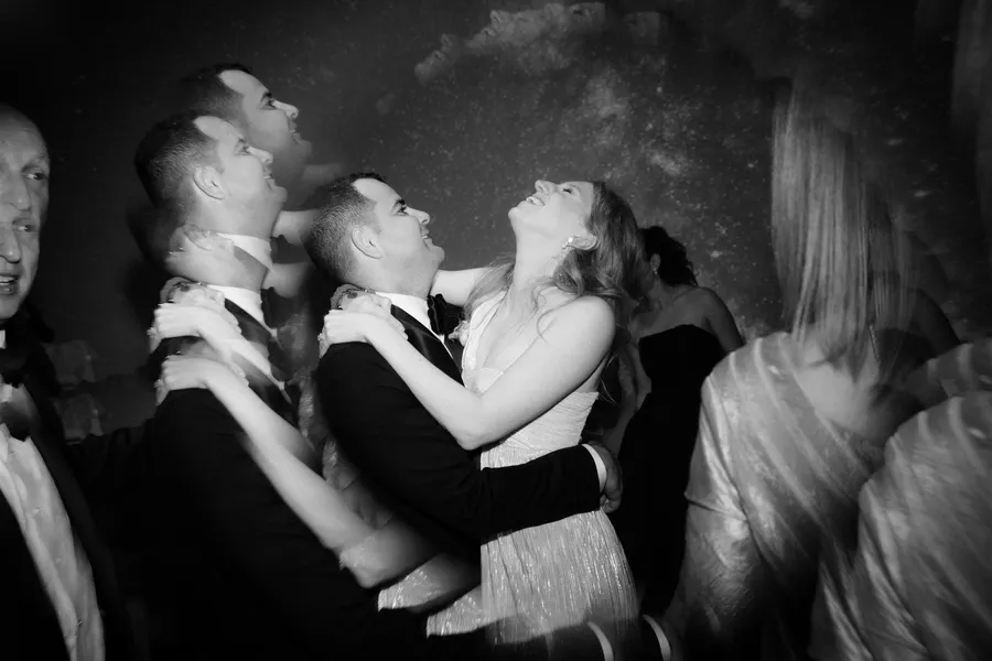 A joyful couple, at their LA wedding reception, embraces and gazes at each other amidst friends in formal attire, with a starry background, all in black and white.