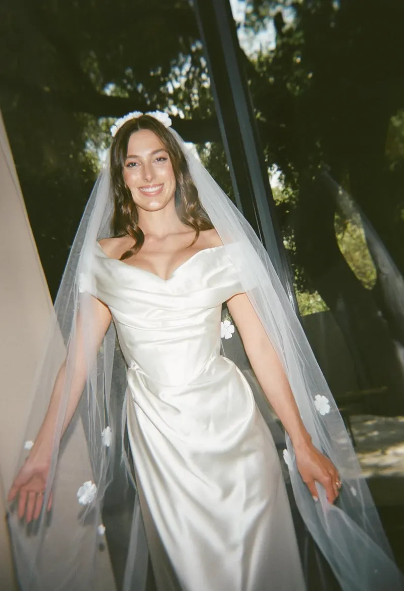 A woman in a satin wedding dress and veil, smiling, standing indoors near a window with greenery outside.