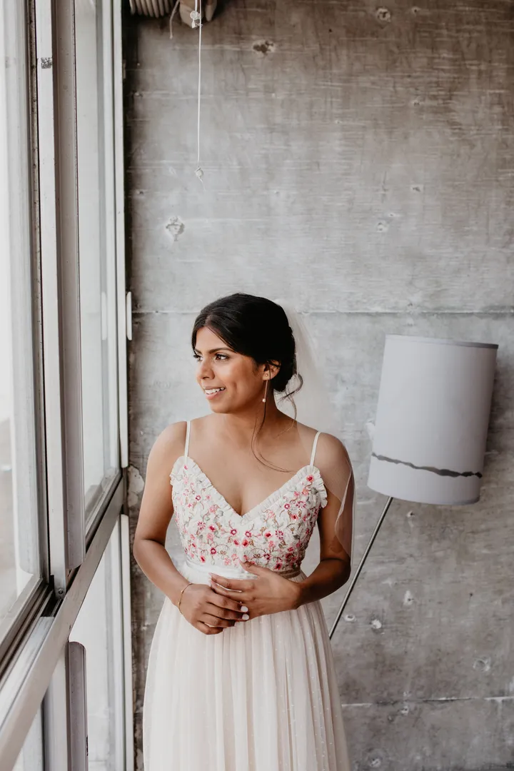 A woman in a white wedding dress with pink embroidery looks out a window, smiling, with a gray concrete wall and a floor lamp in the background.