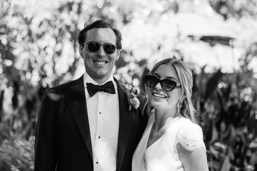 A black and white photo of a smiling man in a tuxedo and a woman in a dress, both wearing sunglasses, standing outdoors with trees in the background.