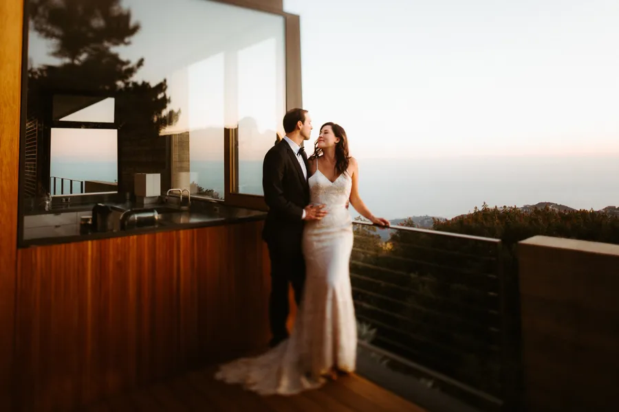 A bride and groom standing on a balcony at sunset, gazing into each other's eyes, with scenic trees and hills in the background.