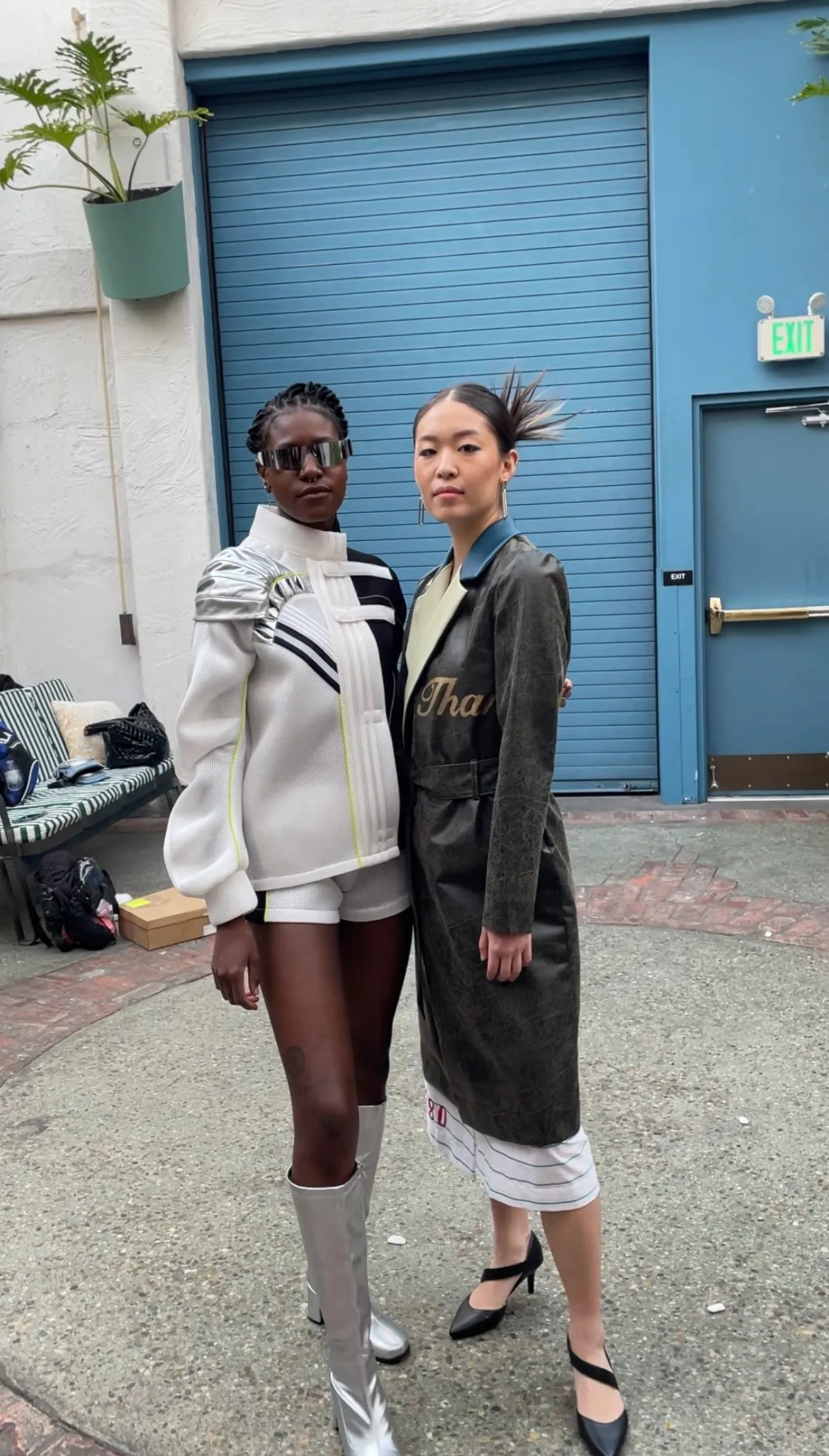 Two women standing together outside, one wearing futuristic sunglasses and a sporty white jacket with matching shorts and tall boots, the other in a fashion-forward black coat, dress, and heels.