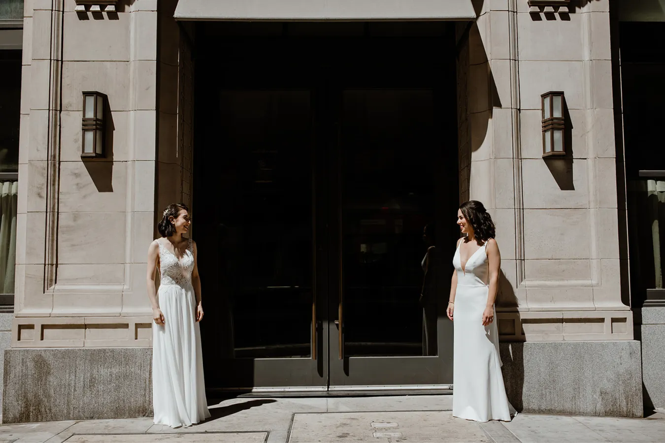 Two women in white dresses standing face to face outside in front of a building with large