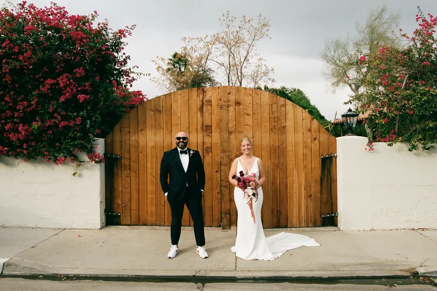 A bride and groom stand in front of a wooden gate surrounded by flowering bushes on either side. The groom is in a tuxedo with sunglasses, and the bride is in a white dress holding a bouquet of flowers. The sky is cloudy.