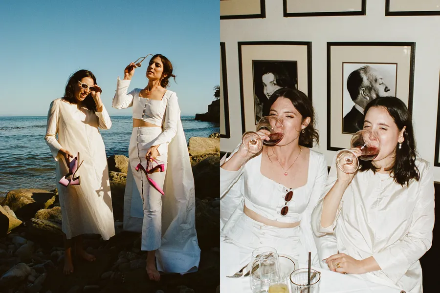 Two women at the beach wearing white clothing and holding pink high heels, one is holding sunglasses, and the other is holding a pair of pink high heels and a glass of wine. The scene is sunny with a clear sky and ocean in the background.