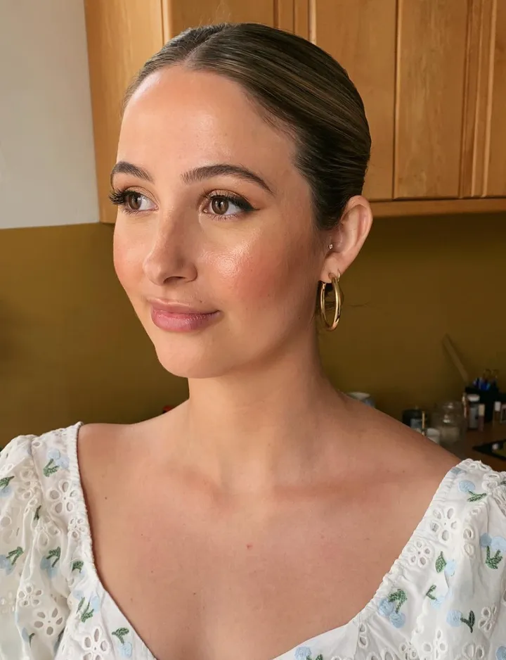 A woman with brown hair tied back, wearing gold earrings and a white blouse with eyelet and floral embroidery, looking to the side, in a room with wooden cabinets in the background.