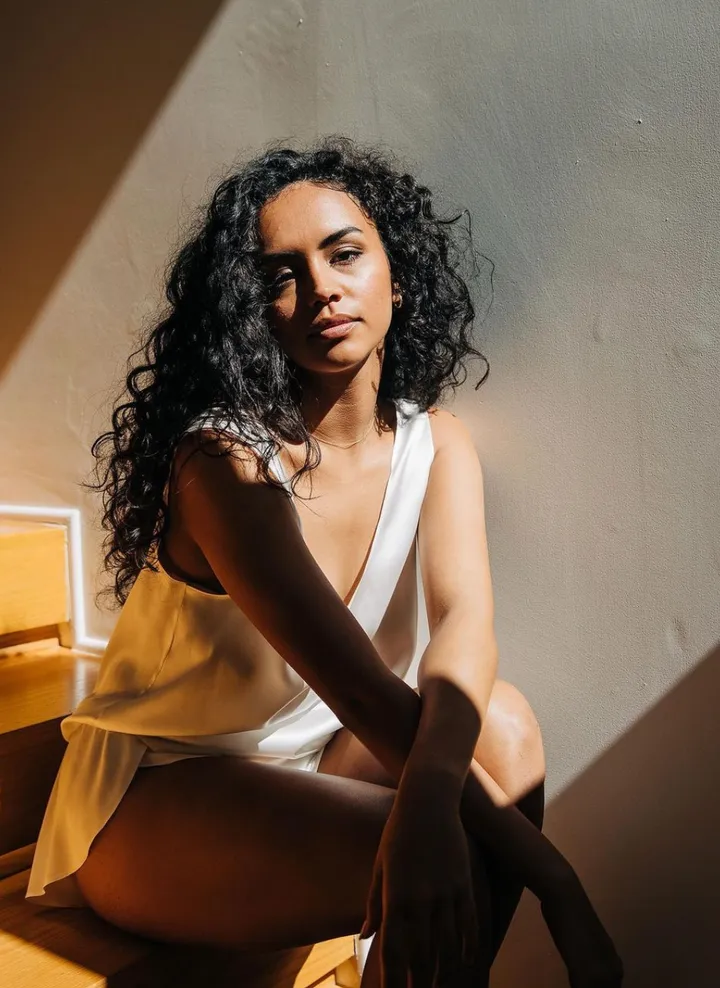 A woman with curly dark hair and light skin sitting on a wooden bench, wearing a white sleeveless top in a sunlit room with a plain wall behind her.