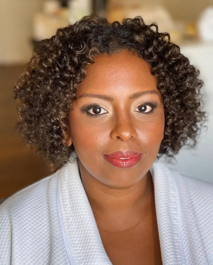 A woman with dark skin and curly hair, wearing makeup with red lipstick, dressed in a robe, getting ready for her red carpet Grammys event in los angeles.