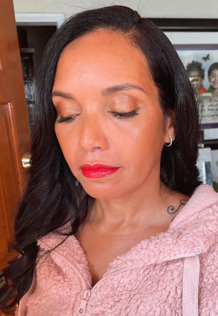 A woman with long dark hair and tan skin, wearing a pink fleece hoodie, has makeup with gold eyeshadow and red lipstick, and is wearing small hoop earrings. Her eyes are closed, and she is indoors.