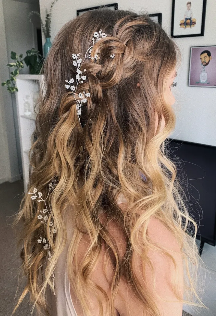A woman with long, wavy blonde hair decorated with a white floral hairpiece, sitting indoors with framed artwork on the wall behind her.