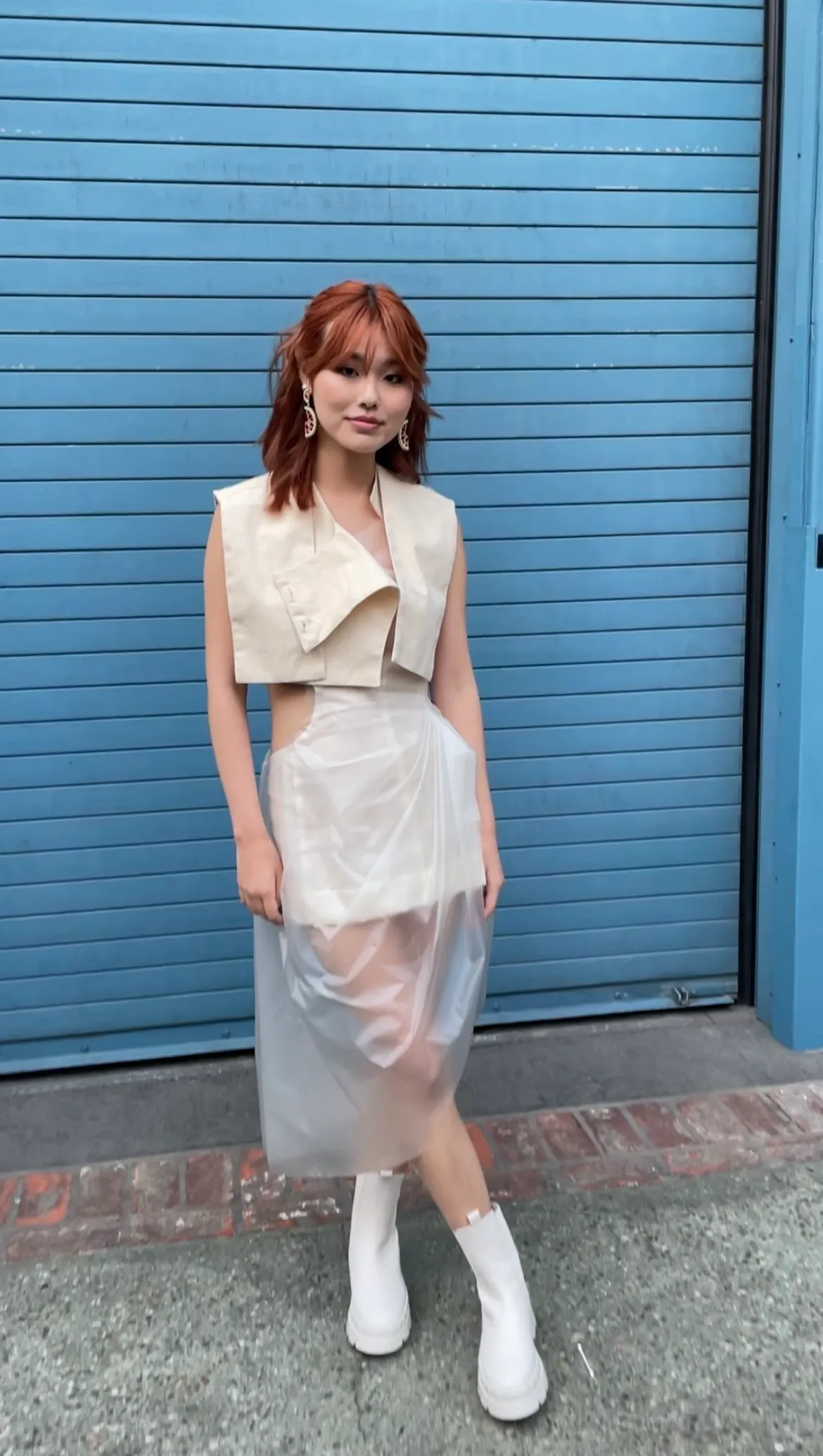 A woman with shoulder-length red hair and earrings stands in front of a blue roll-up garage door, wearing a unique cream-colored dress with cutouts and white boots.