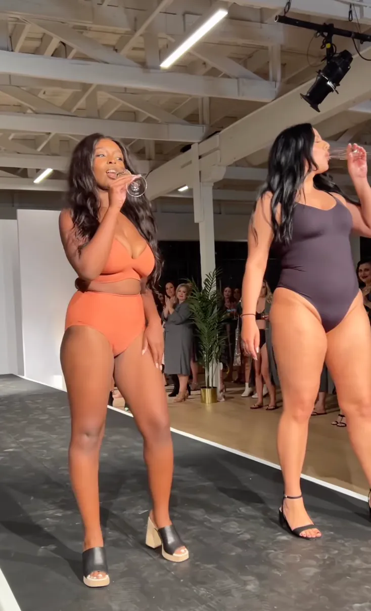Women in swimwear walking on runway at a fashion show, with audience in the background.