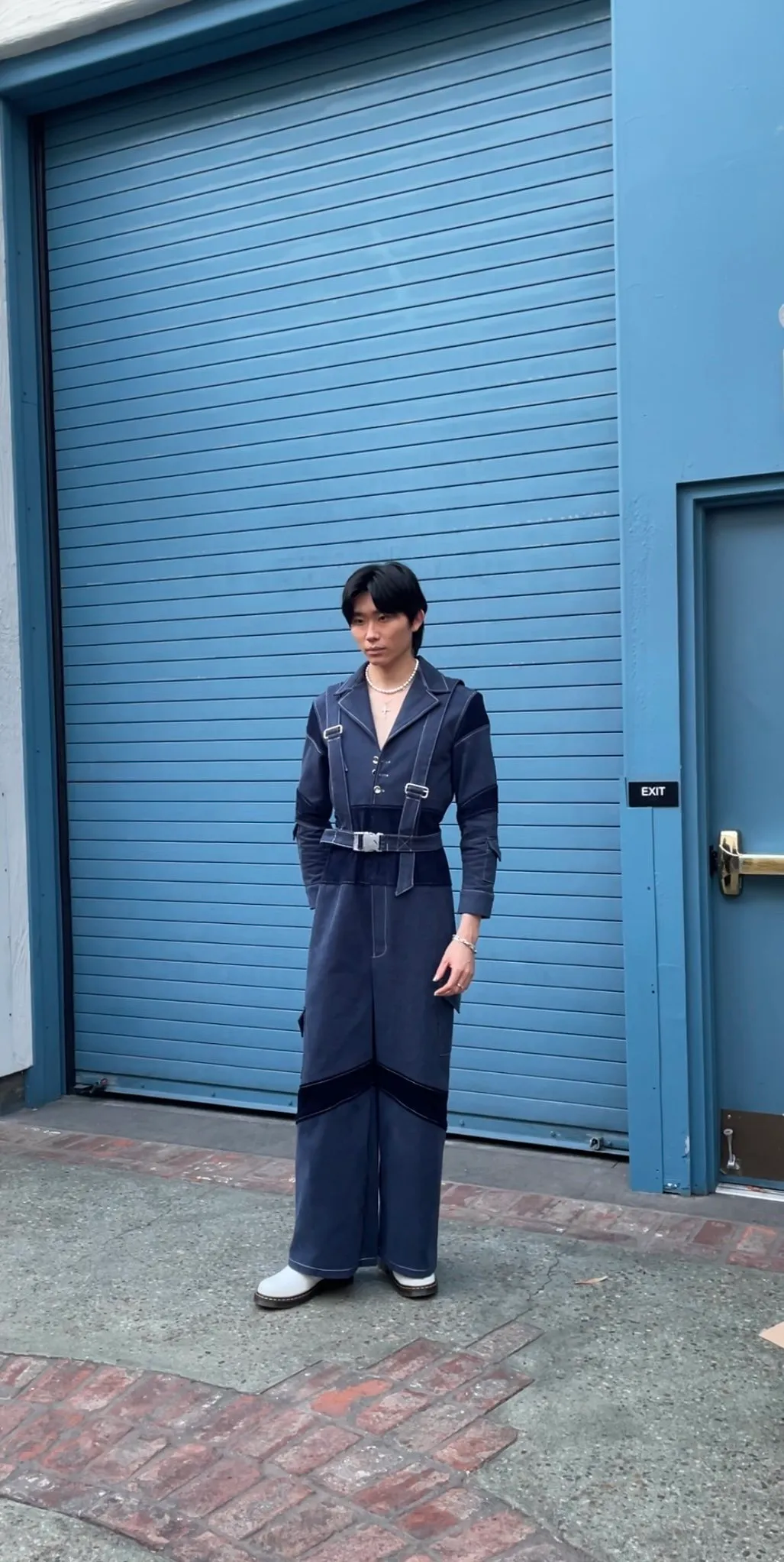 A young man with black hair standing in front of a blue roller door and blue wall, wearing a dark blue jumpsuit and white shoes, with a necklace and bracelet.