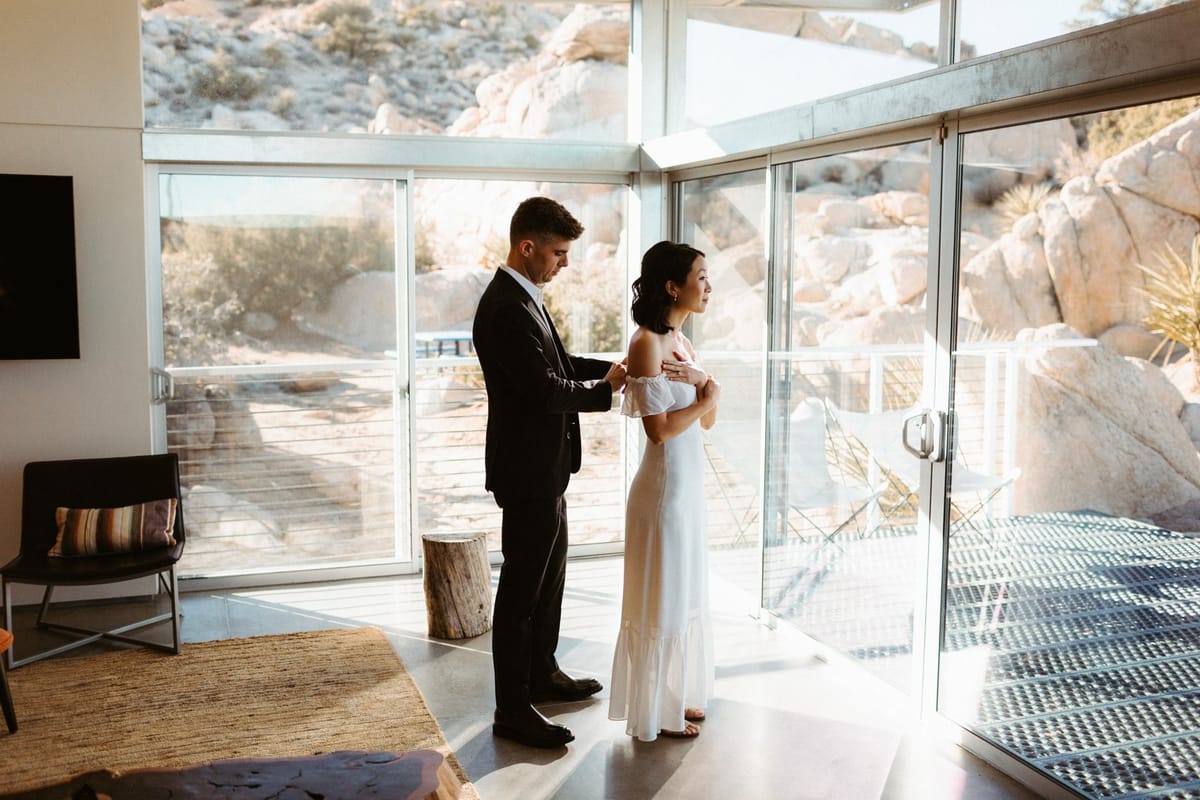 groom zips bride into her dress while getting ready for their intimate elopement in joshua tree